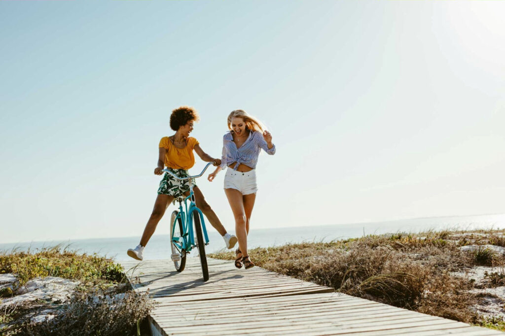 a woman joyfully bikes along a coastal boardwalk as her friend runs beside her, embracing self-care