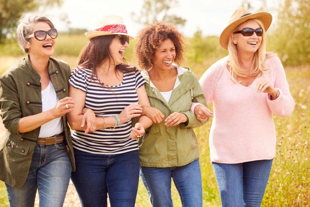 Four high income women with linked arms smiling and laughing while walking down a trail